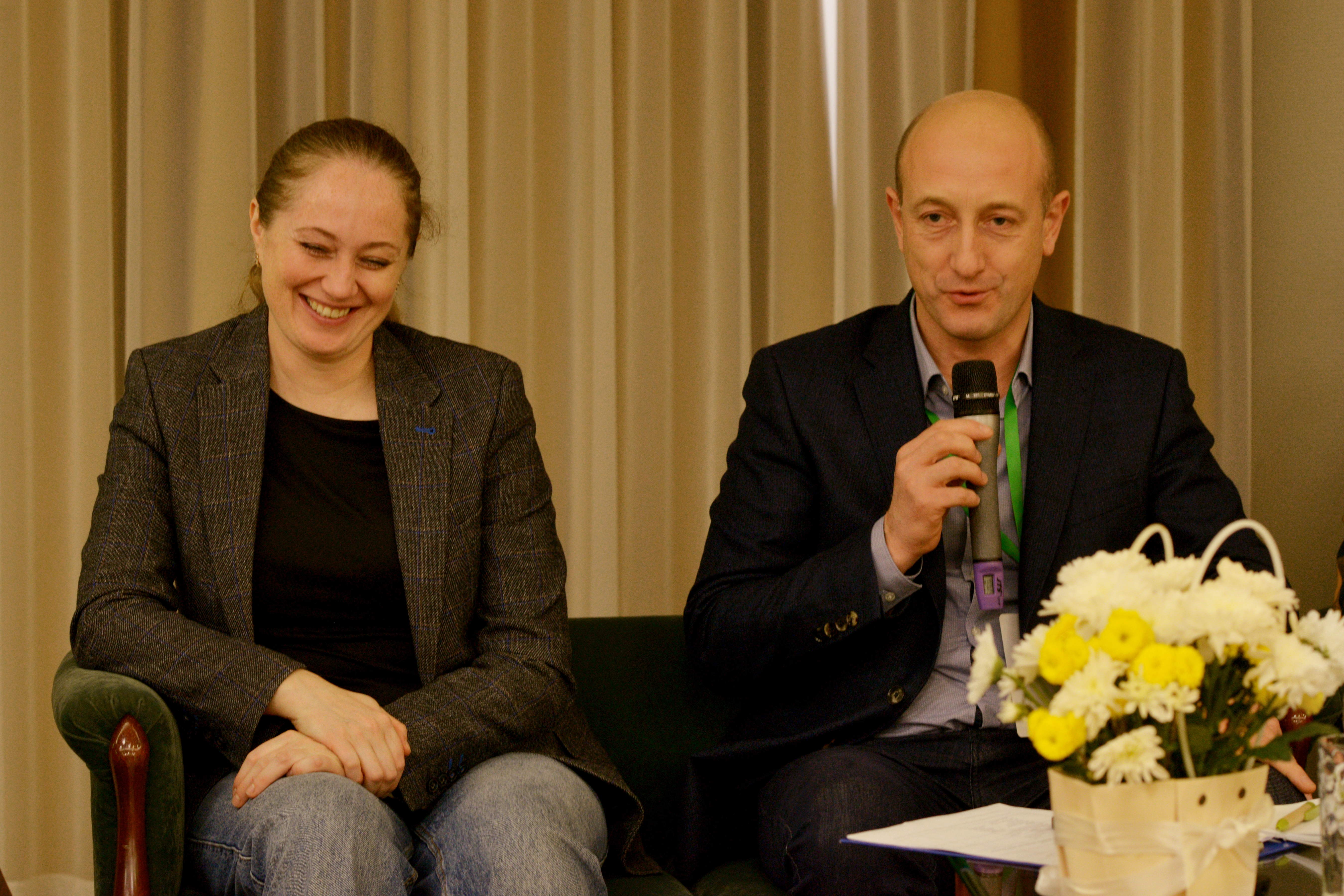 Двоє людей сидят на стільцях, один говорить у мікрофон, пред ними столик на якому стоять квіти