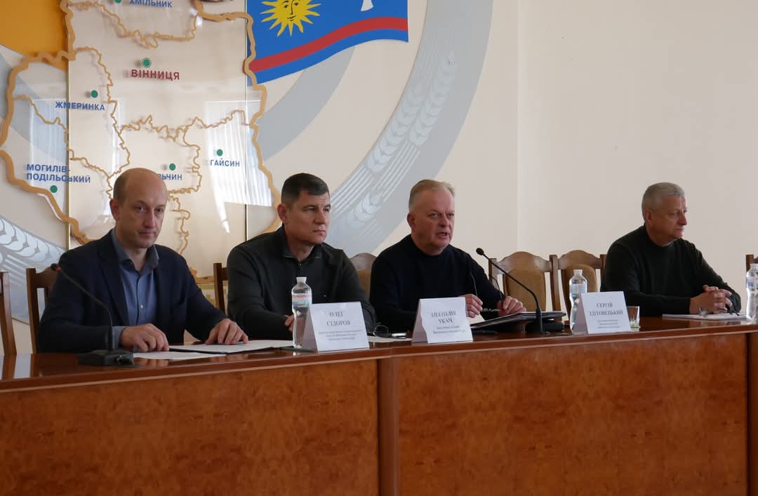Учасники наради сидить за столом перед ними документи, мікрофон та таблички з іменами