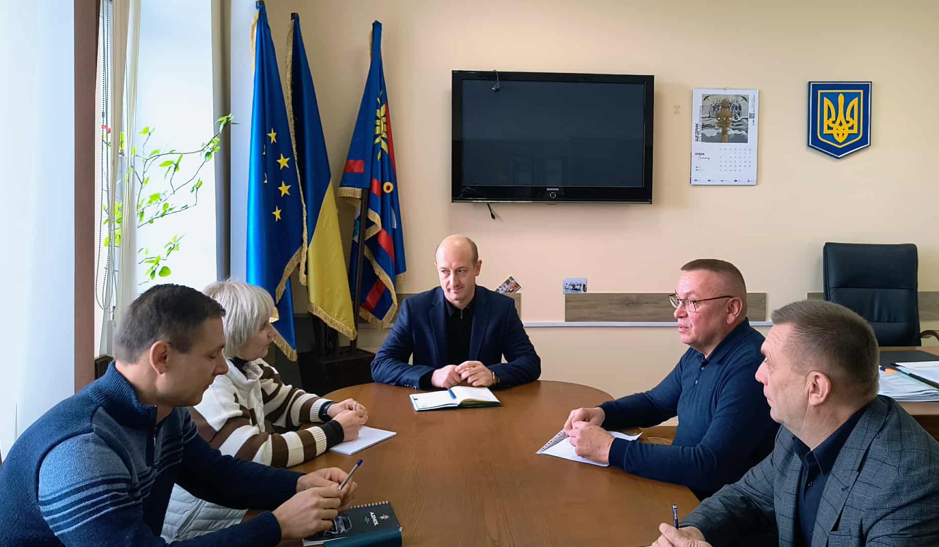 В кабінеті за робочим  столом сидят столом сидят учасники зустрічі, перед ними  документи та записники