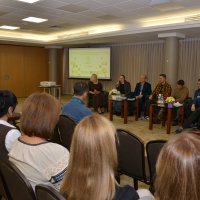 В залі сидить багато людей, одна жінка говорить у мікрофон