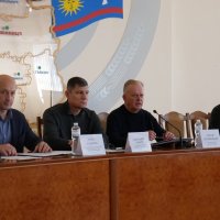Учасники наради сидить за столом перед ними документи, мікрофон та таблички з іменами