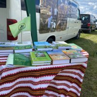 Стіл вкритий вишитою скатертиною на ньому лежать книжки та прапорець з логотипом, позаду стоять автівки учасників заходу перед ними лежать документи